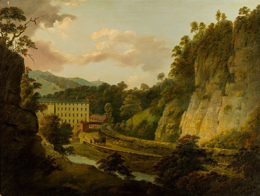 A large mill building sits beside a river at the base of steep, rocky cliffs, surrounded by trees and hills under a partly cloudy sky.