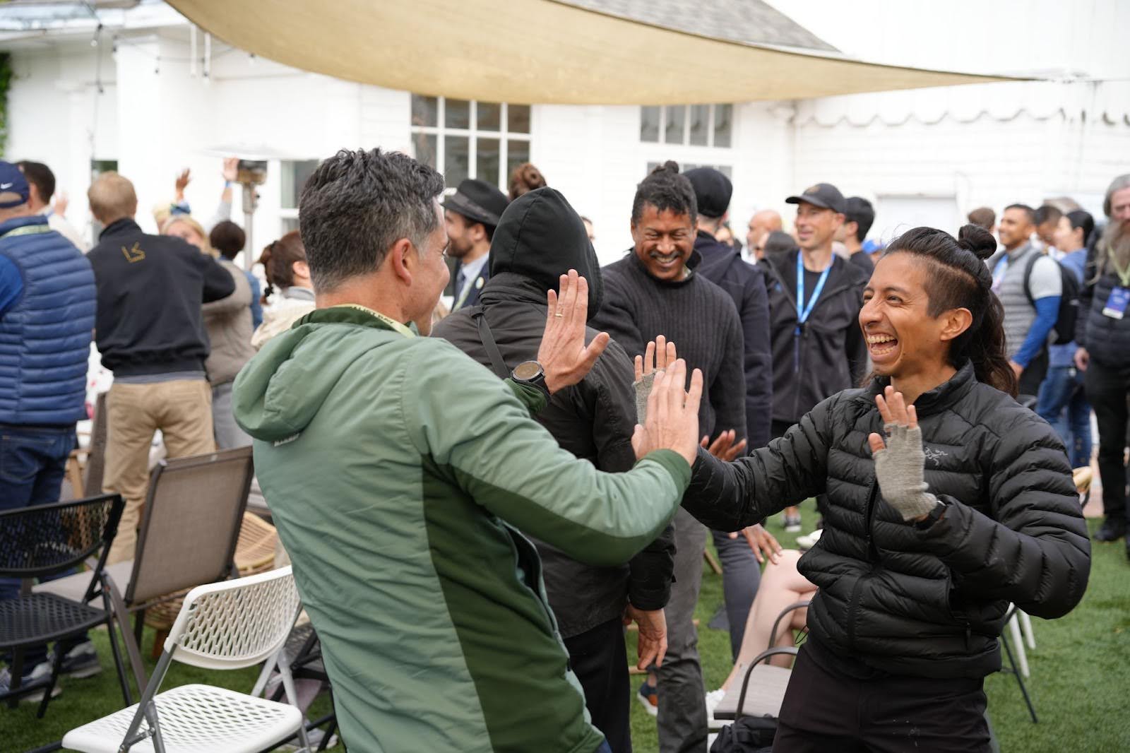 Two people face each other and high-five, smiling, in an outdoor gathering with others standing and chatting in the background.