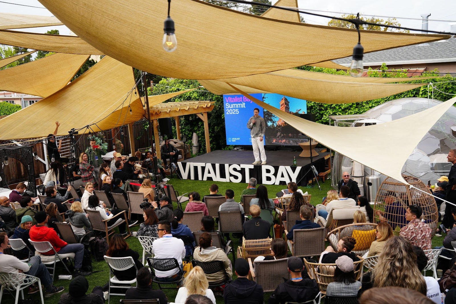 A speaker stands on stage addressing a seated outdoor audience at the Vitalist Bay Summit 2025, with shade sails and greenery in the background.