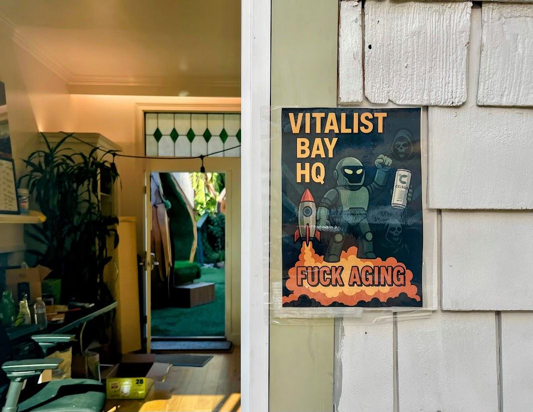 Poster on a wall reads “Vitalist Bay HQ Fuck Aging” with cartoon figures, a rocket, and lab items; an open door reveals a room with plants and green decor inside.