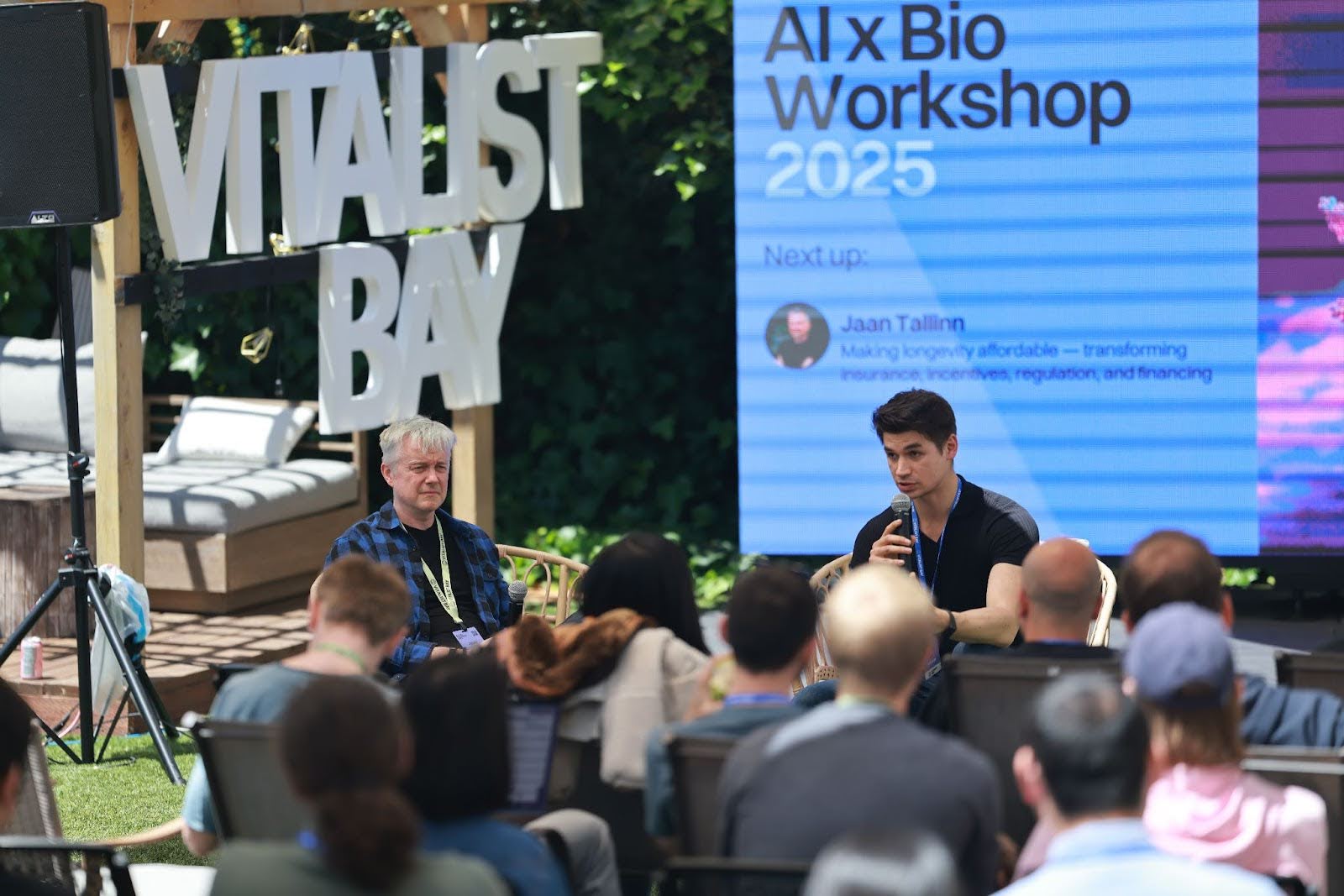 Two speakers address an audience outdoors at the Vitalist Bay AI x Bio Workshop 2025, with a large event sign and presentation screen in the background.
