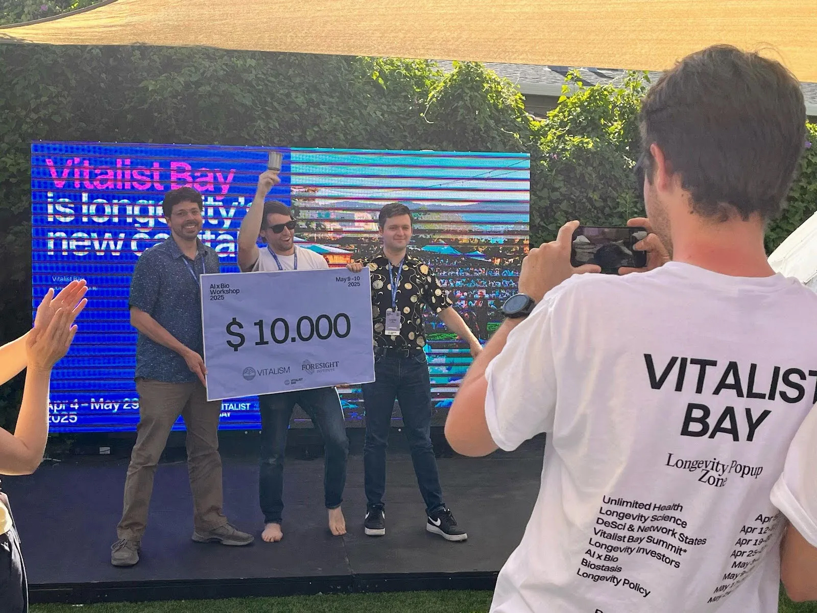 Three men from Vitalist Bay pose on stage holding a $10,000 check as another person takes their photo; a digital sign and greenery are in the background.