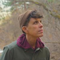 A person wearing a brown beanie and green jacket looks thoughtfully to the side outdoors in a wooded area.