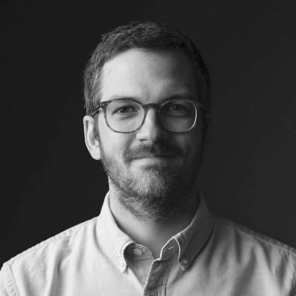 A person with short hair, a beard, and glasses, wearing a button-up shirt, smiles slightly against a dark background.