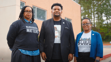 Three people stand outside a brick building, facing forward. The woman on the left wears a "Black Health Matters" shirt, the man in the center wears a blazer, and the woman on the right wears glasses and a blue blazer.
