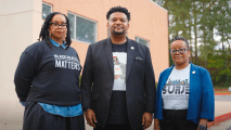 Three people stand outside a brick building, facing forward. The woman on the left wears a "Black Health Matters" shirt, the man in the center wears a blazer, and the woman on the right wears glasses and a blue blazer.
