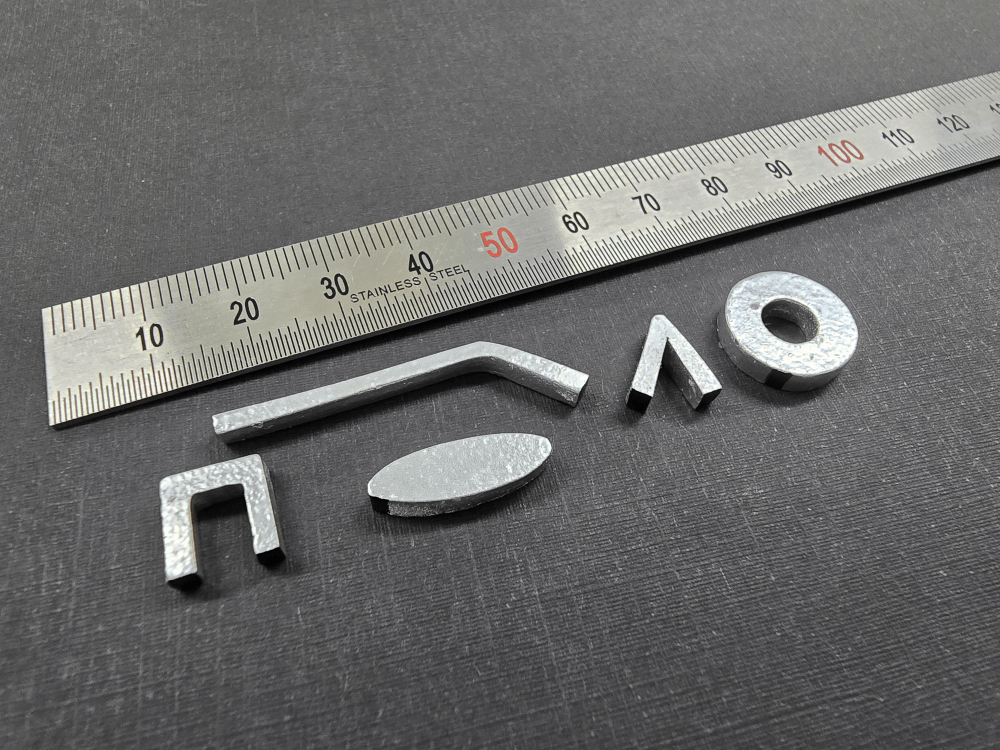 A sleek stainless steel ruler lies alongside five metallic pieces, meticulously arranged in intriguing shapes on a dark surface, reminiscent of avant-garde wearables.